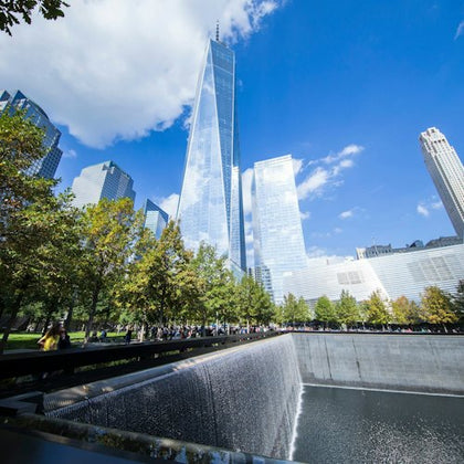 A large reflective skyscraper towers over a memorial fountain surrounded by trees on a sunny day.