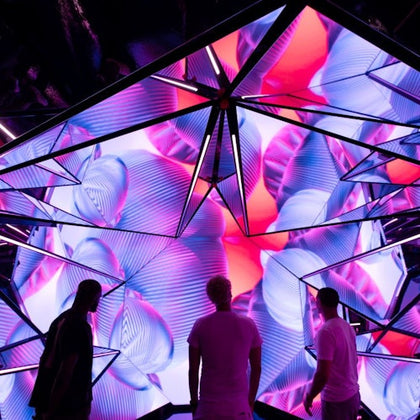 Three people stand in front of a geometric structure displaying vibrant, abstract visuals in purple and pink hues.