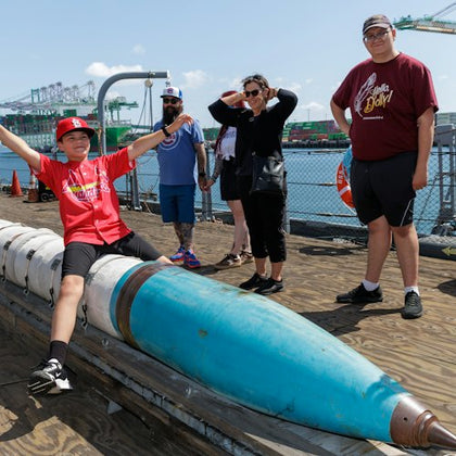 A group of people standing on a pier near a large, blue, cylindrical object, with a harbor and industrial equipment in the background.