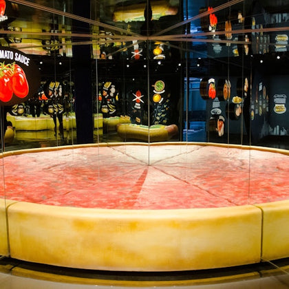 A large, realistic pizza slice sculpture in a mirrored room with various food images and ingredients displayed on the walls.