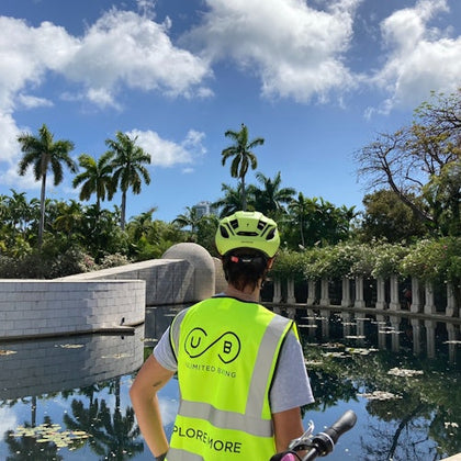 Person in a neon safety vest with "Unlimited Biking" printed on the back, standing by a reflective water feature with palm trees in the background.