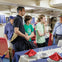 A group of people stand around a dining table set with plates, glasses, and red napkins, inside a ship's interior.