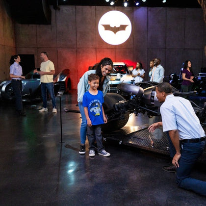 A child and an adult pose for a photo near a Batmobile at an exhibit, with other people and vehicles in the background.