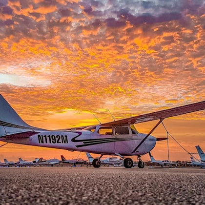 Small airplane parked on a tarmac during a vibrant orange, yellow, and purple sunset with other planes in the background.
