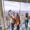 Two people taking a selfie on a glass observation deck with a city skyline, including the Empire State Building, in the background.