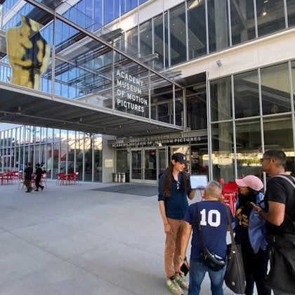 Four people gather outside the entrance of the Academy Museum of Motion Pictures, with a large Oscar statue visible behind glass above.