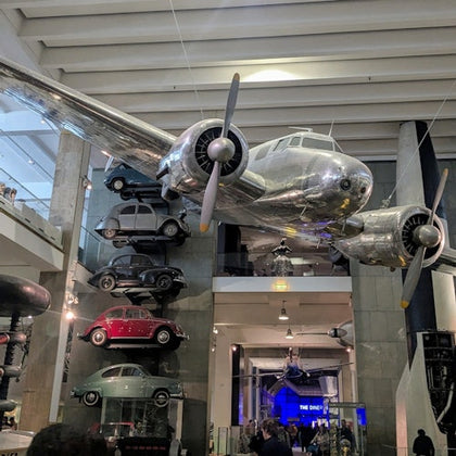An airplane suspended indoors with a display of vintage cars mounted on a wall in a museum setting.
