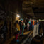 A group of people stands inside a dimly lit tunnel near train tracks, listening to a person speaking next to a display board.