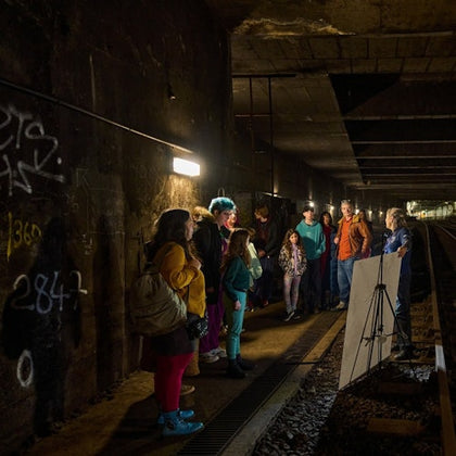 A group of people stands inside a dimly lit tunnel near train tracks, listening to a person speaking next to a display board.