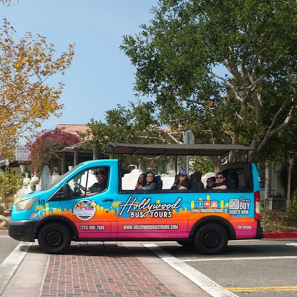A brightly colored tour van labeled 'Hollywood Bus Tours' with passengers, parked on a street near trees and buildings.
