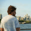 A person with curly hair in a striped shirt stands near a railing overlooking a city skyline across a body of water.