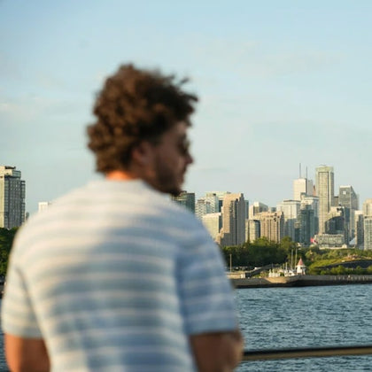 A person with curly hair in a striped shirt stands near a railing overlooking a city skyline across a body of water.