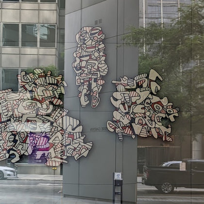 Abstract graffiti-style art displayed on a glass building facade with city reflections and passing cars.