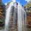 A tall waterfall cascades down a rugged cliff surrounded by lush vegetation under a bright blue sky.