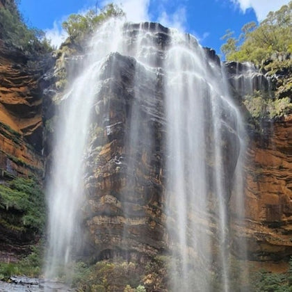 A tall waterfall cascades down a rugged cliff surrounded by lush vegetation under a bright blue sky.