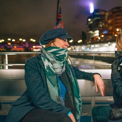 Two people sitting on a boat at night, one wearing a scarf and hat, with city lights blurred in the background.