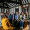 A group of people seated indoors in a room with wooden beams, listening attentively to someone off-frame.