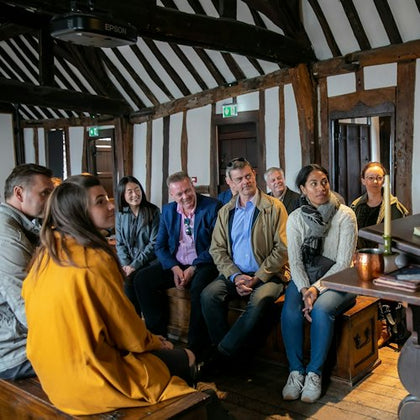 A group of people seated indoors in a room with wooden beams, listening attentively to someone off-frame.