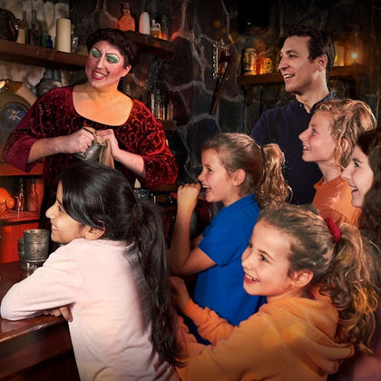 A woman in medieval costume interacts with a smiling man and a group of children in colorful clothing inside a rustic setting.