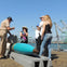 A group of people on a dock, with a uniformed man showing a large, blue torpedo-like object. Cranes are visible in the background.