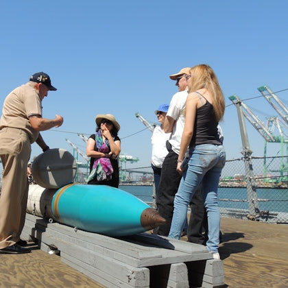 A group of people on a dock, with a uniformed man showing a large, blue torpedo-like object. Cranes are visible in the background.