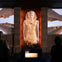 A large Egyptian statue displayed in a museum, with two people observing it, set against a backdrop of temple columns.