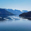 Several speedboats creating wakes on a calm lake, surrounded by forested hills and snow-capped mountains under a clear blue sky.
