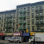 A row of urban buildings with green fire escapes, storefronts below, and vehicles parked on the street in front.