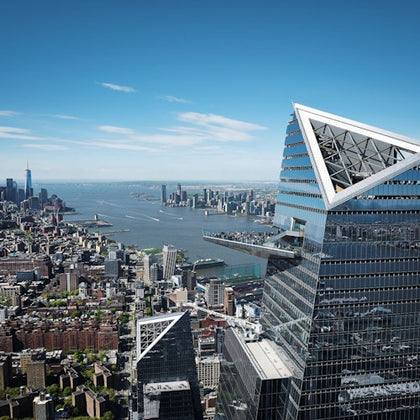 A cityscape featuring modern skyscrapers, including a triangular-topped building, with a river and distant skyline visible.