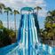 A man and a child in an orange inflatable tube enjoying a pool with water slides and palm trees in the background.