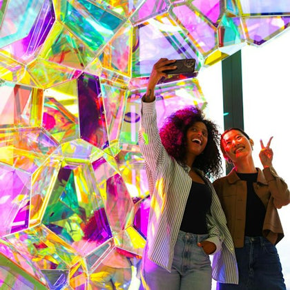 Two people smiling and taking a selfie in front of a colorful, geometric, translucent art installation.