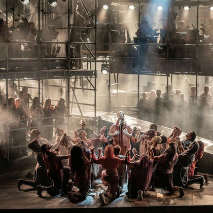 A theatrical performance showing a group of people in dramatic poses on a stage with scaffolding and lighting in the background.