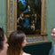 Three people viewing a framed Renaissance painting depicting a woman and children with a naturalistic background in a museum.