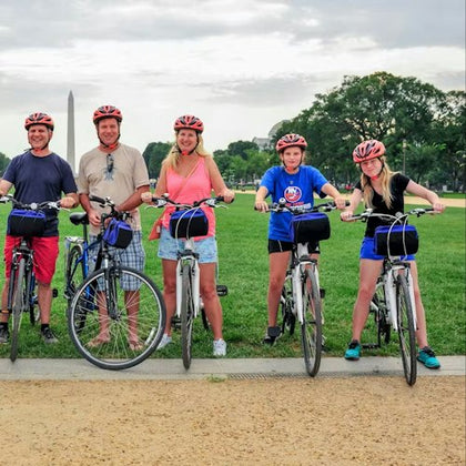 Five people wearing helmets stand with their bicycles on a path in a park, with the Washington Monument visible in the background.