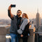 A couple taking a selfie on a rooftop deck at sunset, with a city skyline and the Empire State Building in the background.