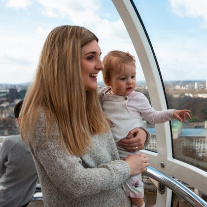 A woman holding a baby inside a large observation wheel pod, smiling and looking out at the cityscape below.