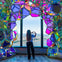 A person stands by a window framed with colorful geometric panels, forming a heart shape with their hands, overlooking a cityscape.