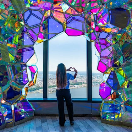 A person stands by a window framed with colorful geometric panels, forming a heart shape with their hands, overlooking a cityscape.