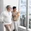 Two men stand by a large window overlooking a cityscape with tall buildings and a river, one pointing out the view.