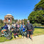 A group of people posing with bicycles in a park near a large domed structure and trees under a clear blue sky.