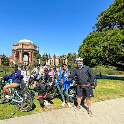 A group of people posing with bicycles in a park near a large domed structure and trees under a clear blue sky.