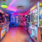 A colorful arcade room with game machines, neon lighting, and a vending machine in the back.