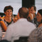 Group of people sitting and talking around a table at an indoor event, with one woman smiling and another man resting his chin on his hand.