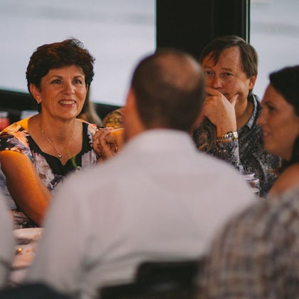 Group of people sitting and talking around a table at an indoor event, with one woman smiling and another man resting his chin on his hand.