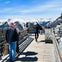 People standing on a wooden observation deck with snowy mountain peaks in the background under a clear blue sky.