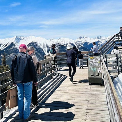 People standing on a wooden observation deck with snowy mountain peaks in the background under a clear blue sky.