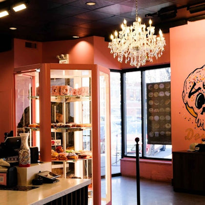 A cozy donut shop with pink walls, pastry display case, chandelier, and a large donut-themed mural near the window.