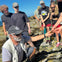 A group of people outdoors participating in a hands-on activity involving seaweed, led by a man wearing a hat and sunglasses.