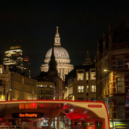 Nighttime cityscape featuring St. Paul's Cathedral dome illuminated, with a red double-decker bus in the foreground.
