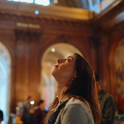 A woman looking upwards in a warmly lit, ornate interior space with arched details and wood paneling.